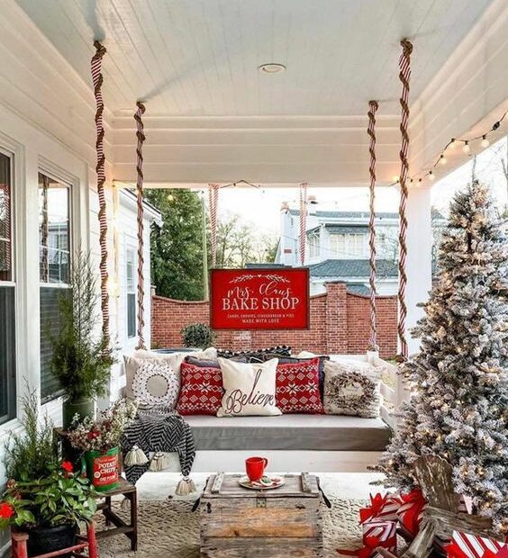 a bright rustic Christmas porch with a flocked Christmas tree with lights mini trees in buckets red pillows and a wooden table 1
